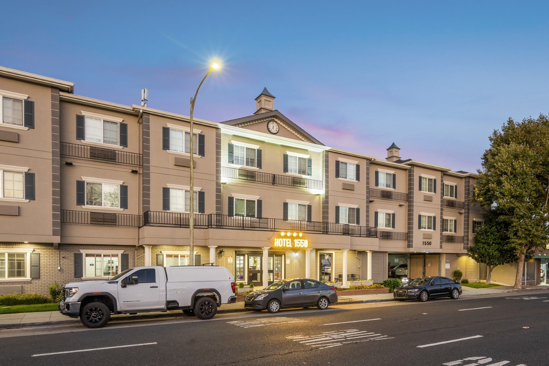 A three-story beige hotel at twilight with a white truck parked in front on a street.