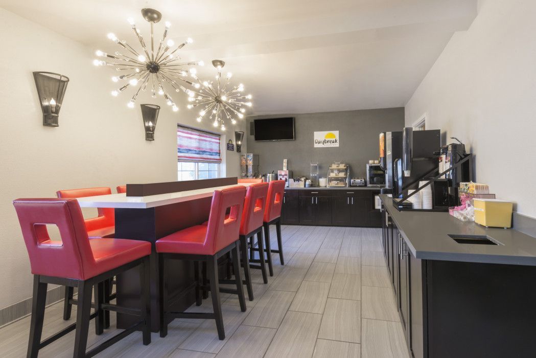 A modern hotel breakfast area with red bar stools at a central counter, pendant lighting, and a coffee station.