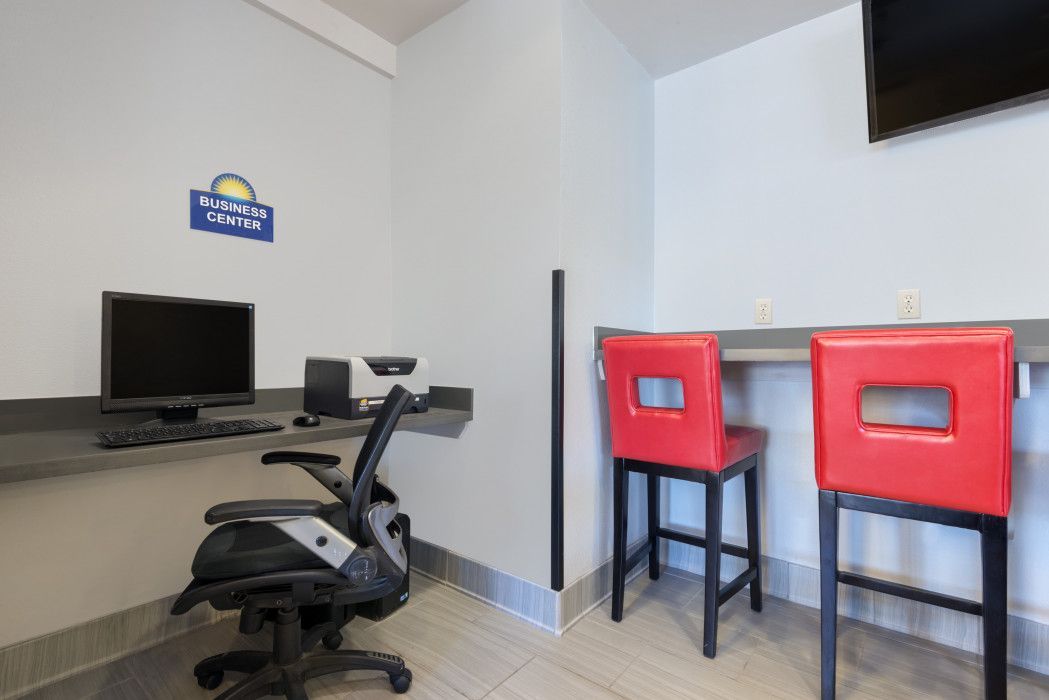 A business center with a desktop computer, printer, office chair, and two red bar stools at a counter under a TV.