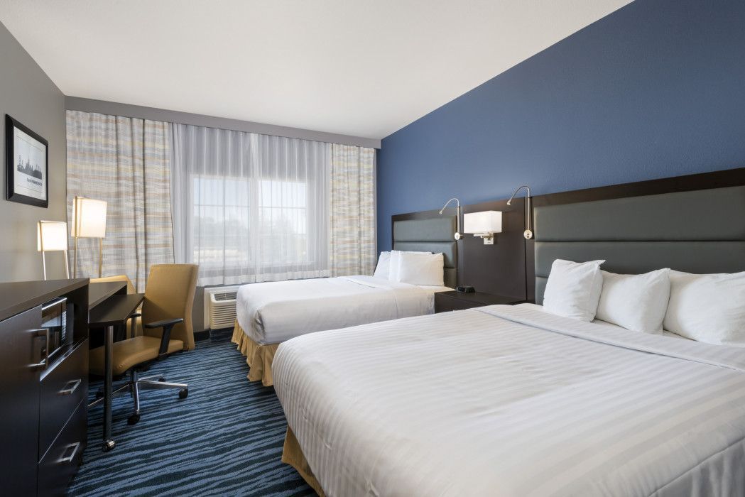 A hotel room with two queen beds, a desk and chair, and a blue accent wall with modern lighting.
