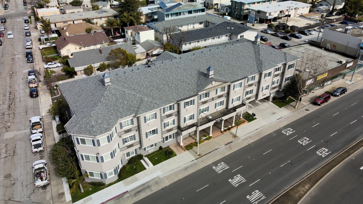 An aerial view of a multi-story apartment complex positioned between a residential street and a multi-lane road.