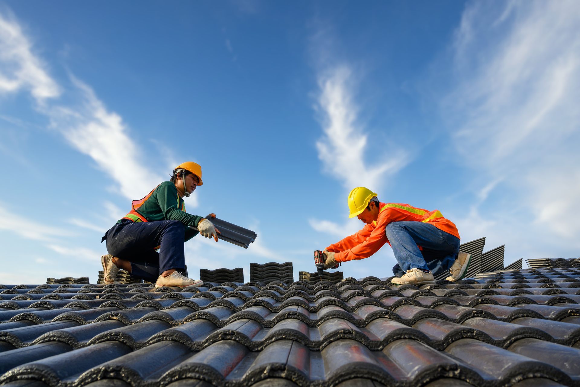Two roofers installing dark tiles on a roof, blue sky background.
