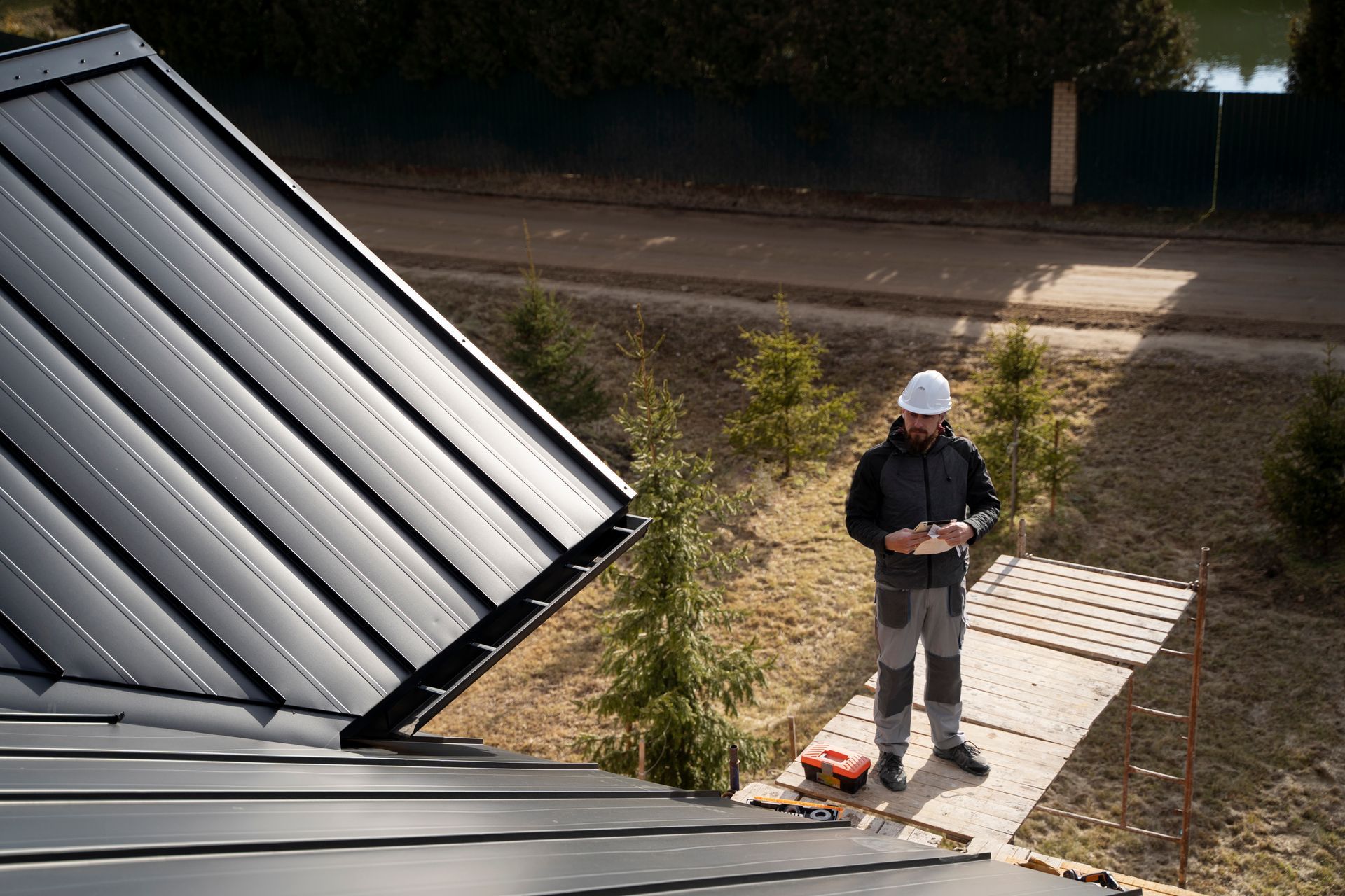 Man in hard hat stands near a black metal roof, holding a tablet; red toolbox beside him.