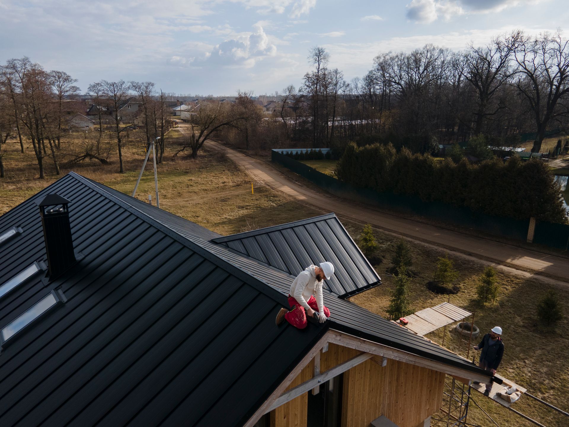 Workers installing black metal roof on a house in a rural setting; sunny day.