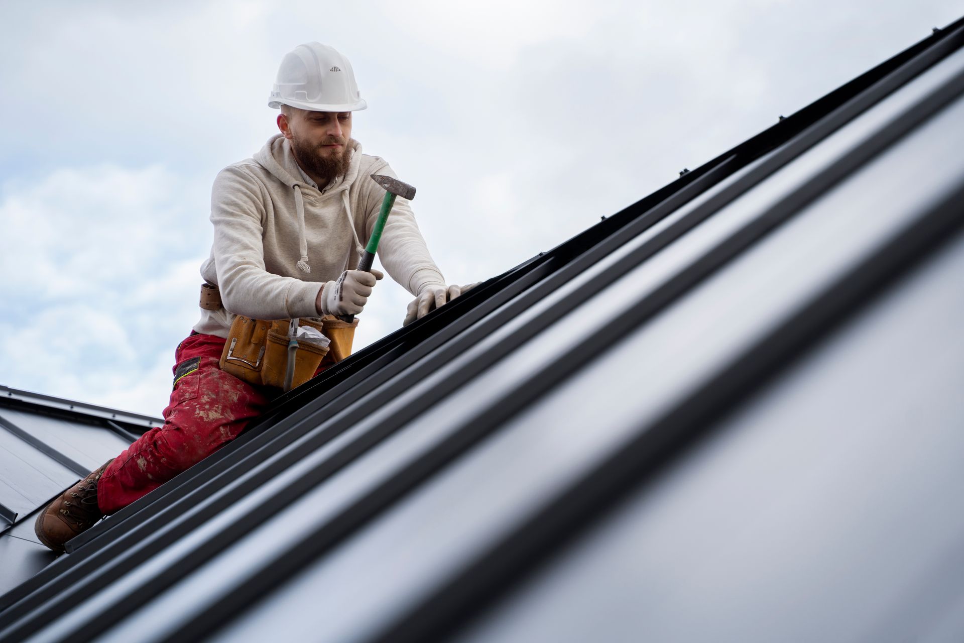 Roofer in hard hat hammering on a metal roof under cloudy sky.