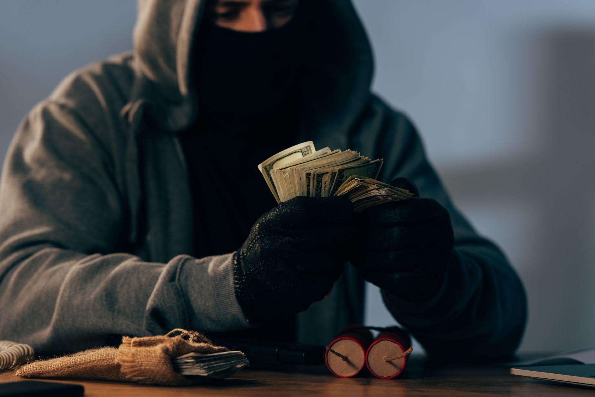 Person in hood and mask holding money, dynamite on table.