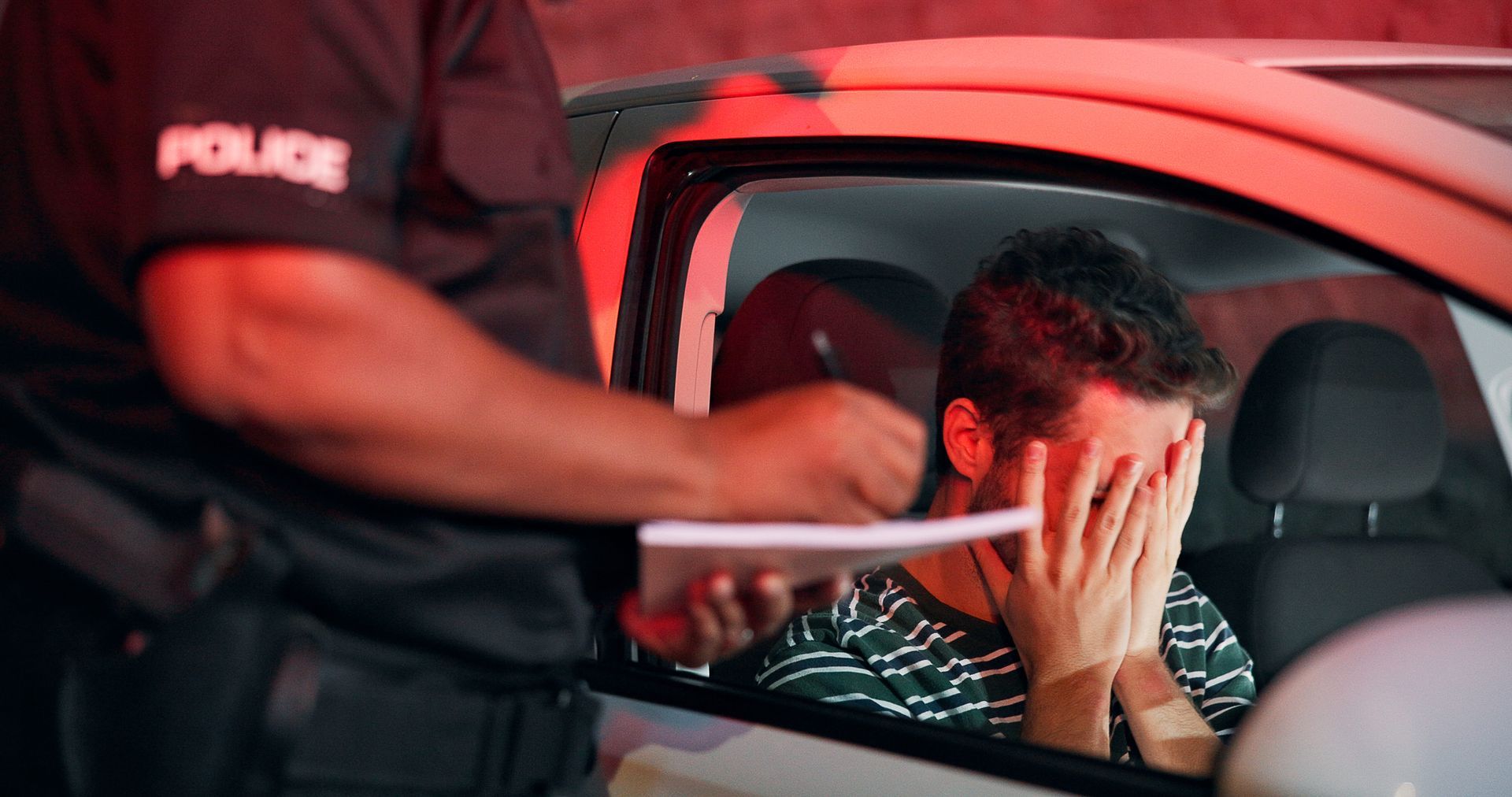 Police officer detaining a man in a car, the man covers his face under red emergency lights.