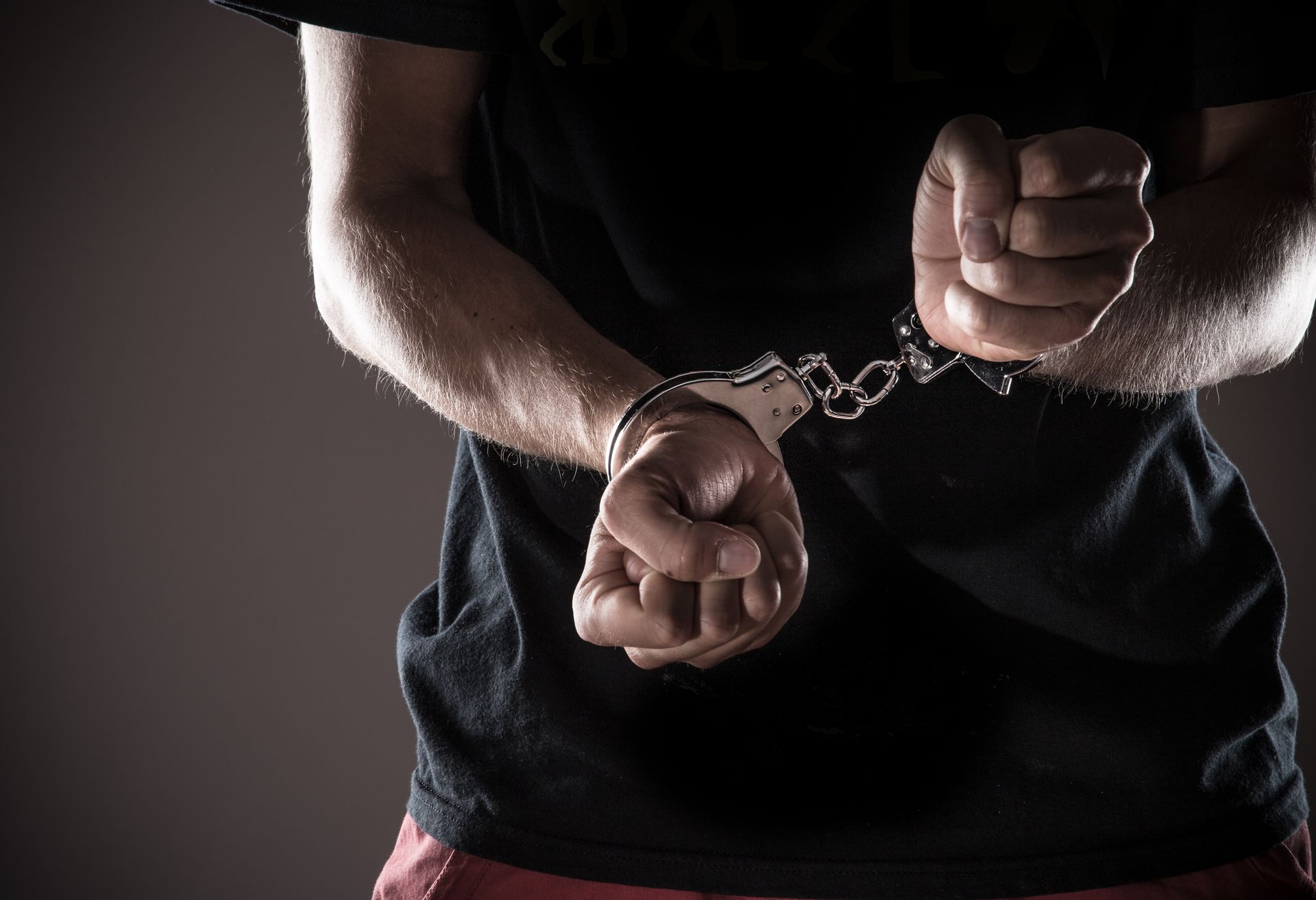 Person in handcuffs, dark shirt, hands clasped, gray background.