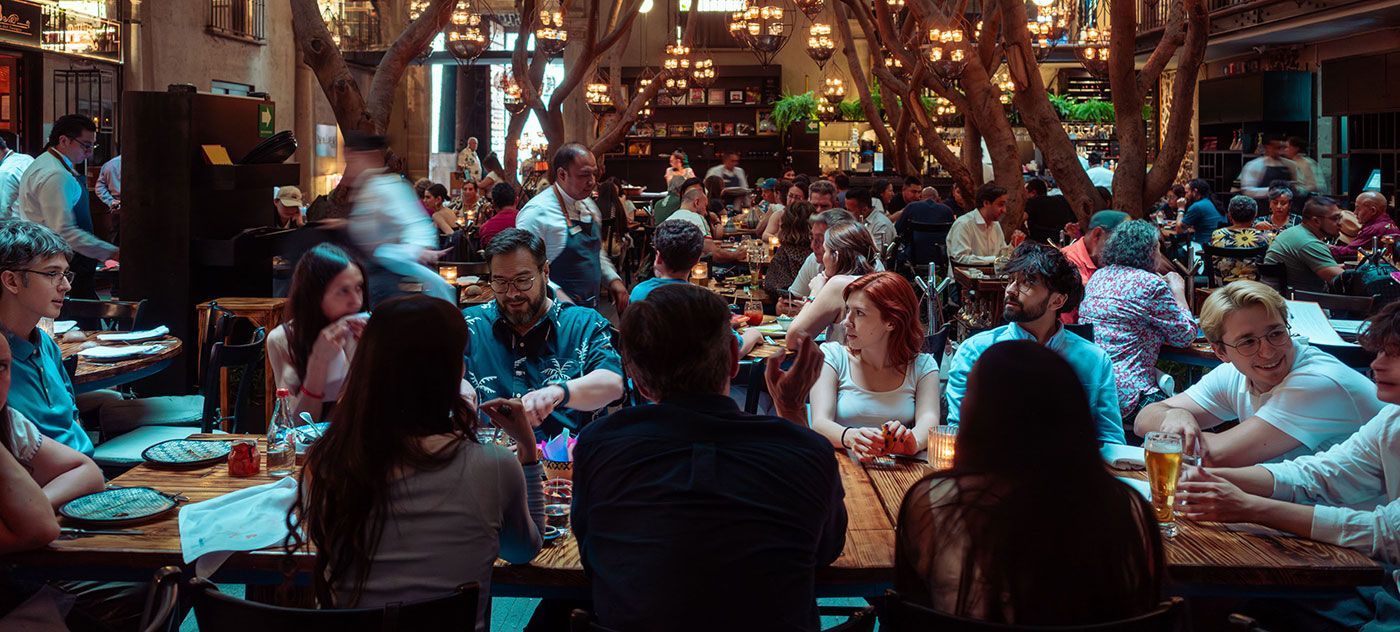 Comedor de restaurante abarrotado con gente comiendo en largas mesas bajo cálidas luces colgantes.