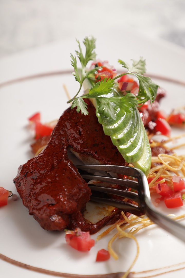 Primer plano de un plato de carne con salsa, hierbas, tomates picados y un tenedor sobre un plato blanco.