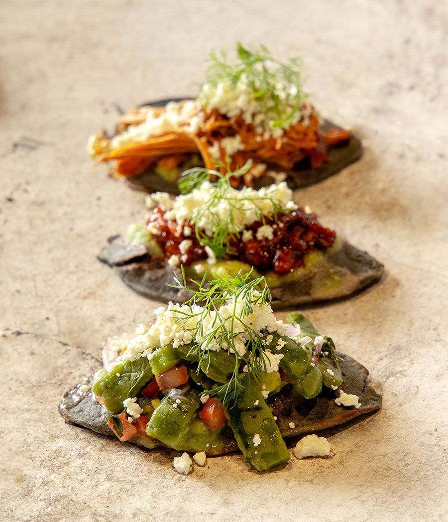 Tres tacos adornados sobre una superficie de piedra, cubiertos con hierbas, frijoles y verduras.