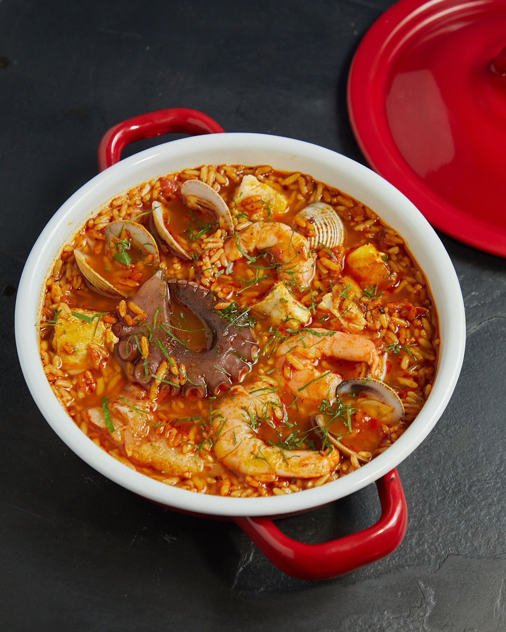 Una olla roja con guiso de camarones sobre una encimera oscura, con una tapa roja cerca.