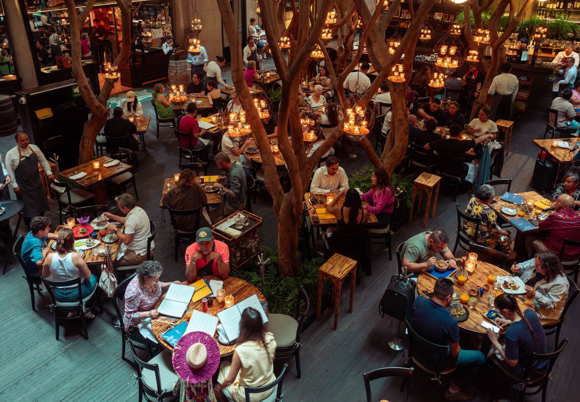 Cafetería interior abarrotada de gente comiendo en mesas bajo cálidas luces colgantes y vigas de madera.