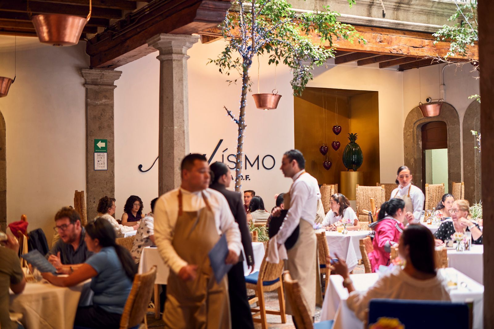 Gente cenando en un animado restaurante con patio interior, iluminación cálida y plantas colgantes.