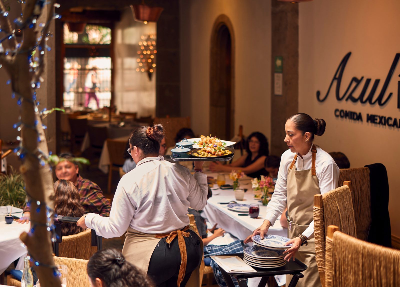 Camareros llevando bandejas en un restaurante con iluminación cálida, comensales sentados y un letrero de