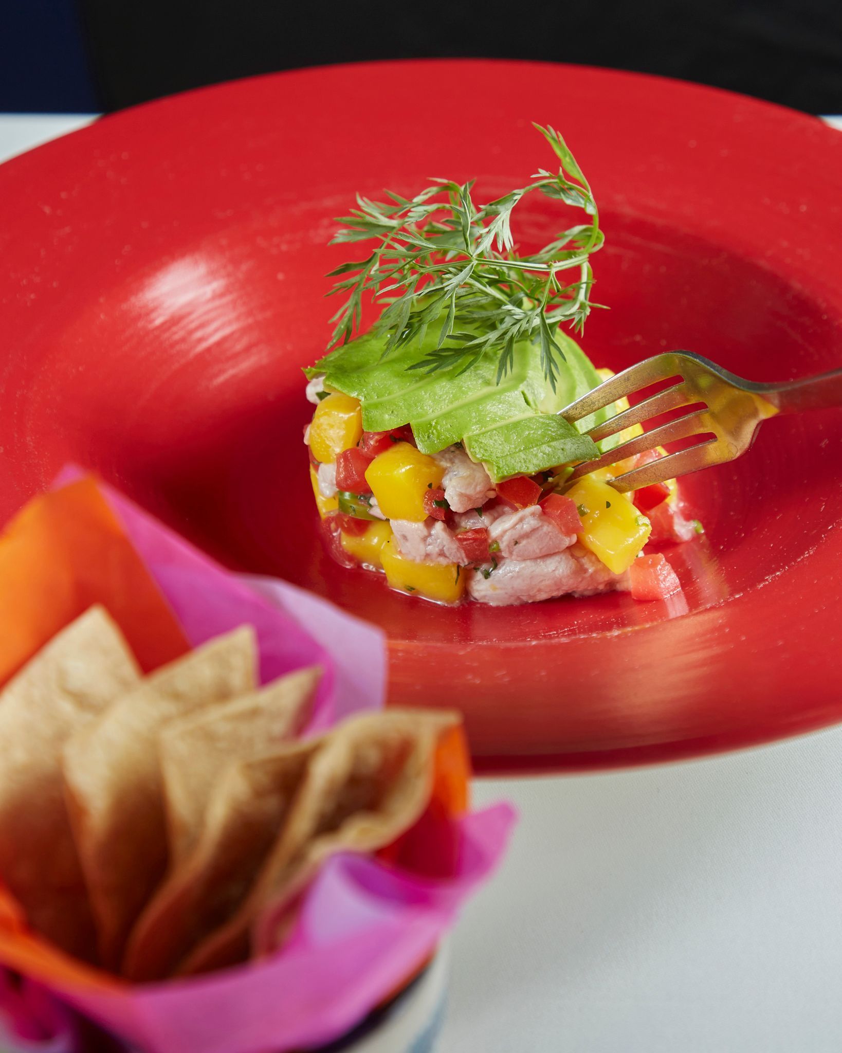 Aperitivo colorido servido en un tazón rojo, cubierto con hierbas, acompañado de totopos y guarnición rosa.