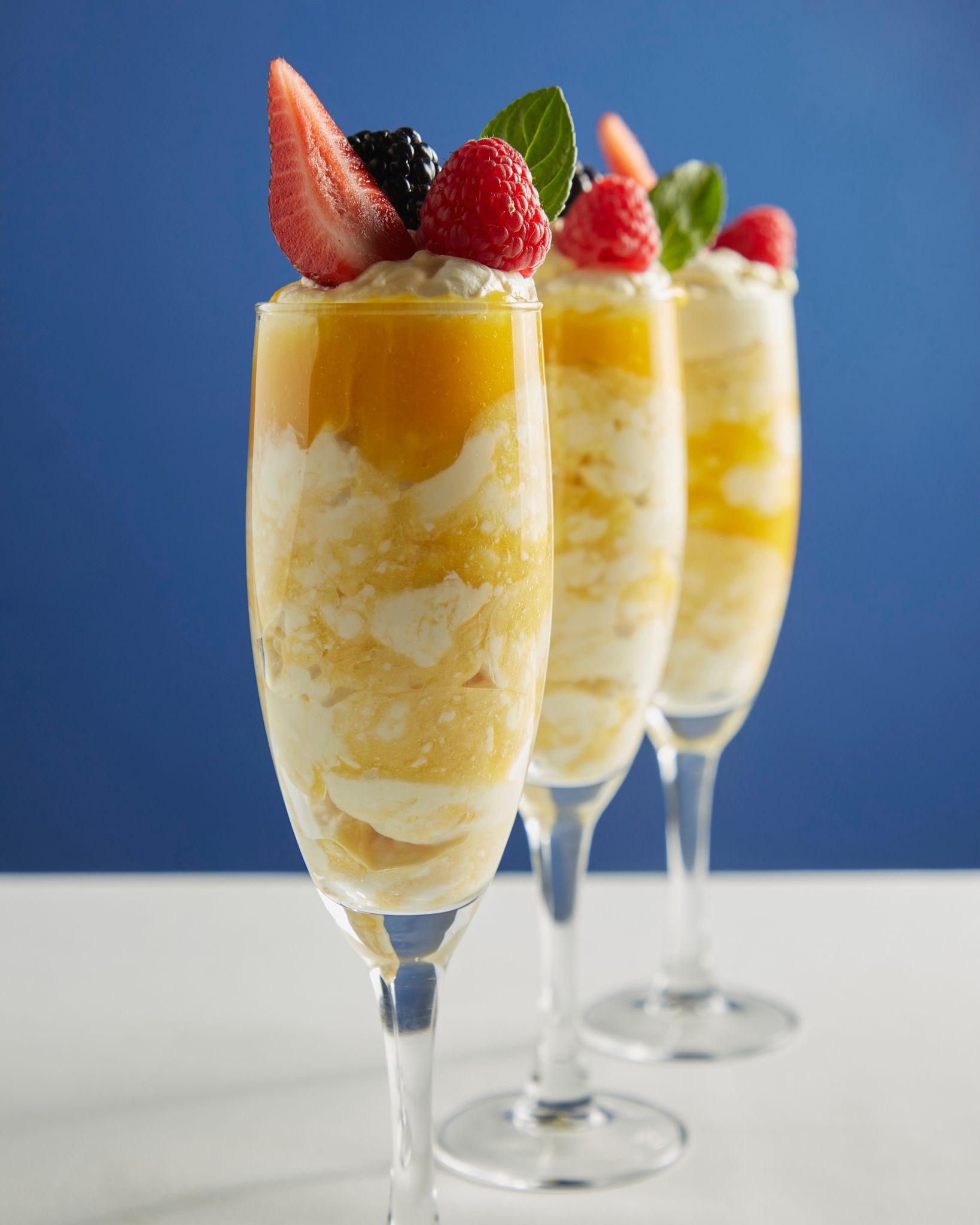 Tres copas de champán rellenas de postre de mango en capas, coronadas con fresas sobre un fondo azul.