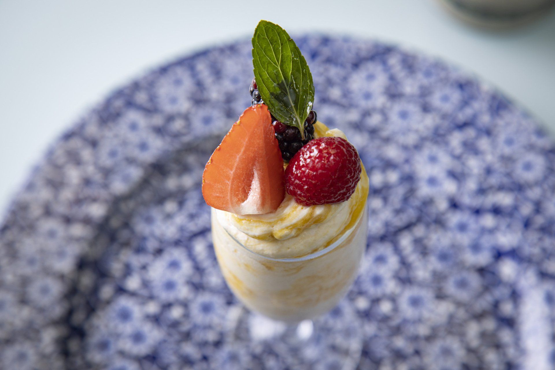 Vaso de postre decorado con fresa, rodaja de naranja y hoja de menta sobre un plato con estampado azul.