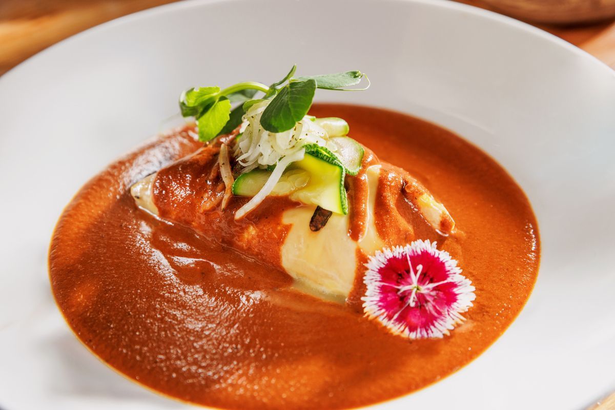 Sopa roja en un tazón blanco, decorada con hierbas, verduras y una flor comestible rosa.