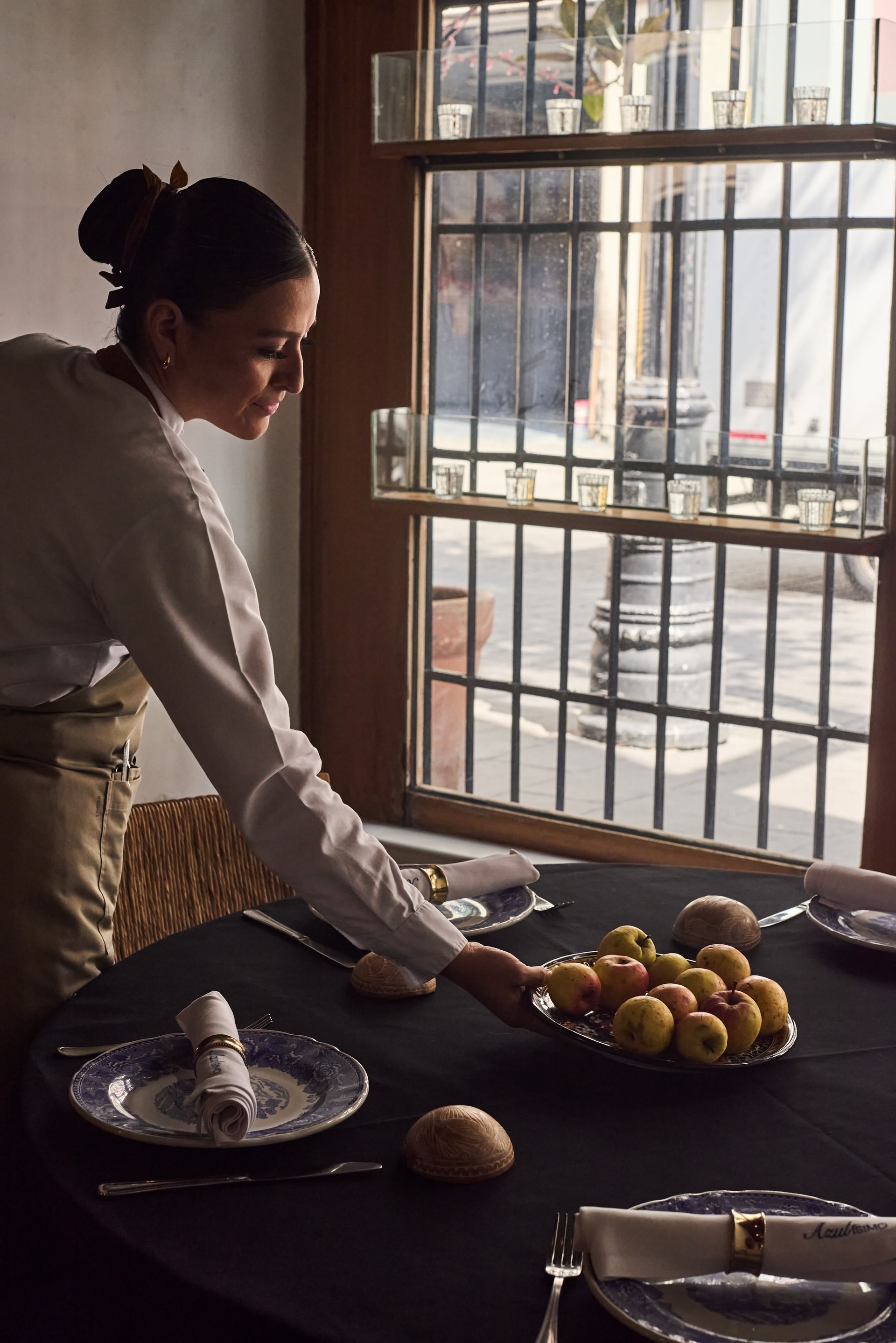 Mujer poniendo la mesa junto a una ventana, con platos, fruta y tazas en una habitación iluminada por el sol.