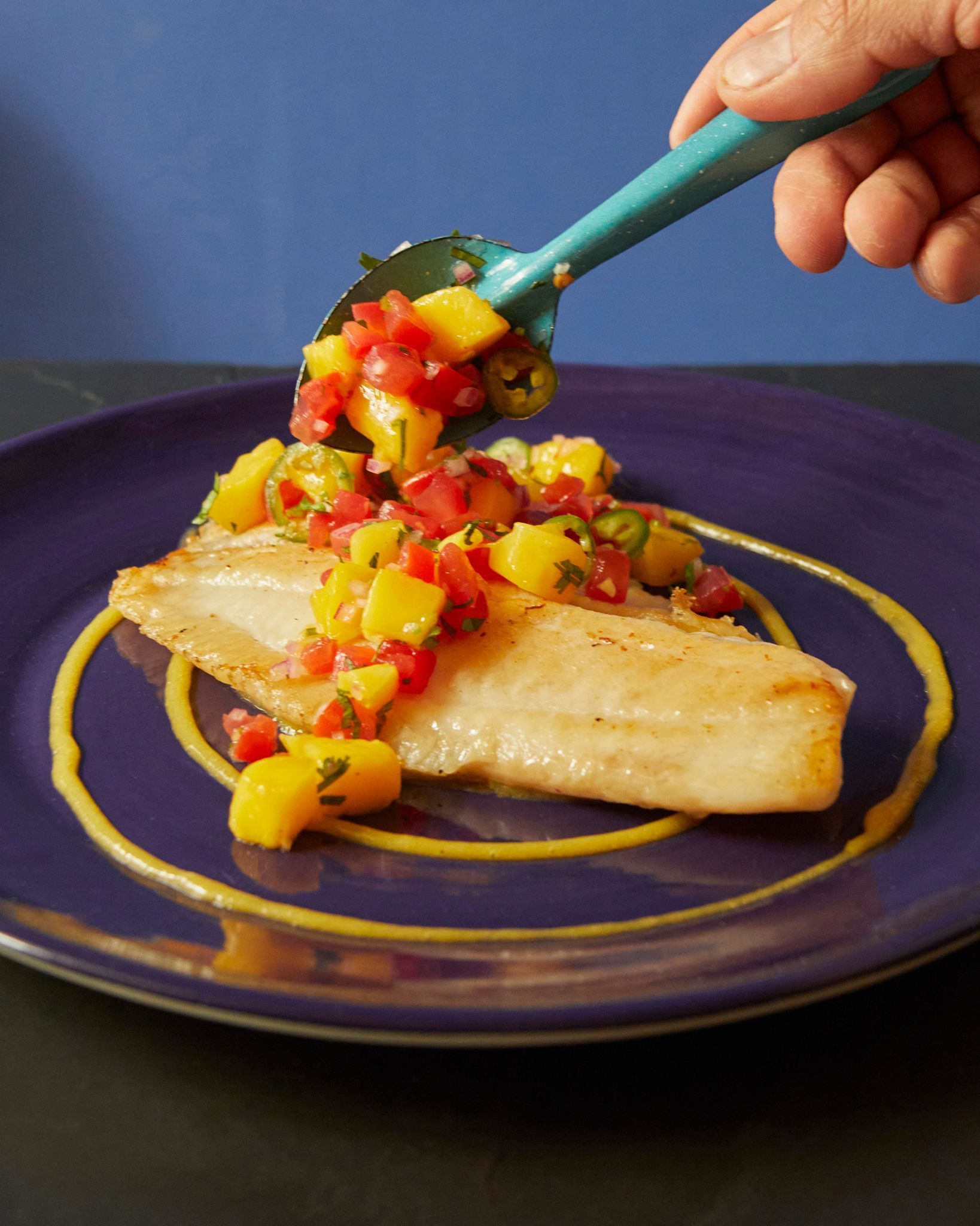 Una mano coloca salsa colorida sobre un filete de pescado a la parrilla en un plato morado.