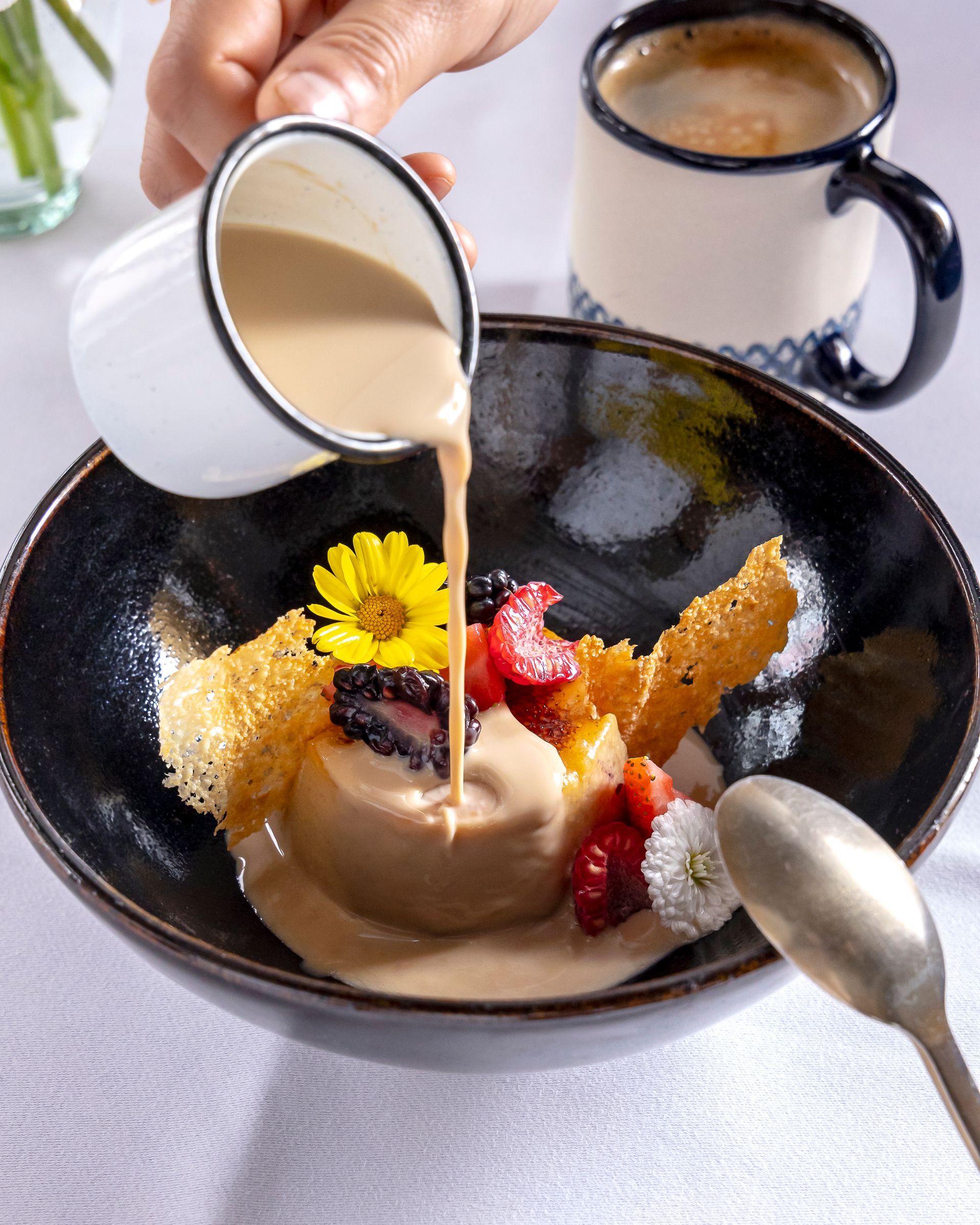 Postre en un tazón negro con salsa de crema, bayas, flor amarilla y dos tazas sobre una mesa.