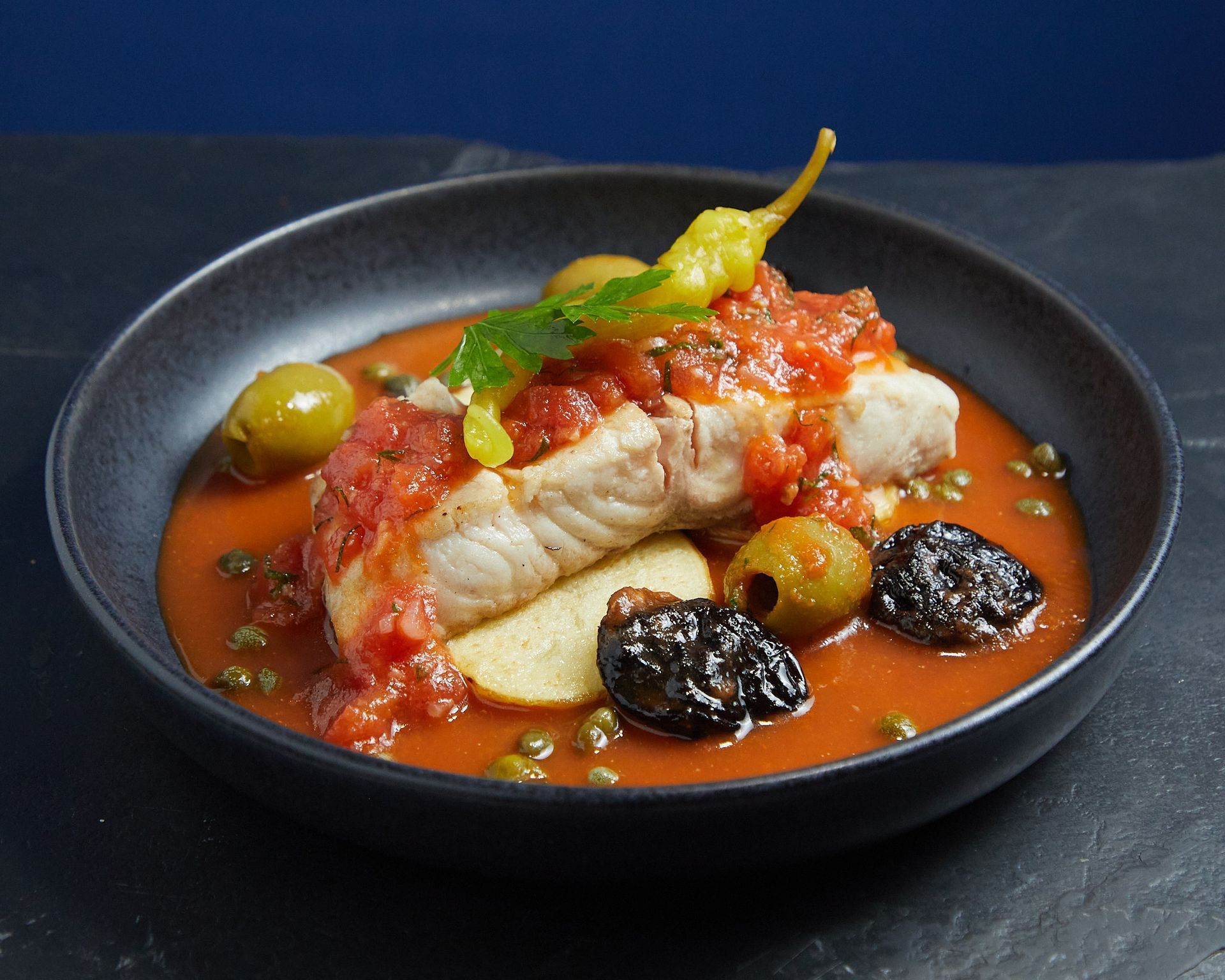 Pescado emplatado en salsa roja con aceitunas y guarnición en un plato oscuro.