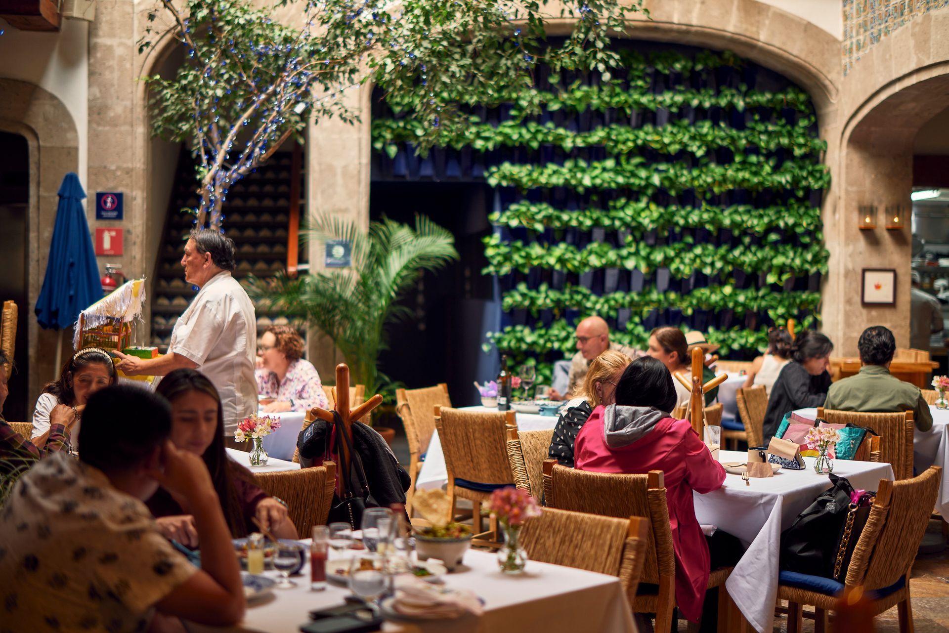 Patio de restaurante al aire libre con comensales sentados en mesas junto a una exuberante pared verde bajo arcos de piedra.