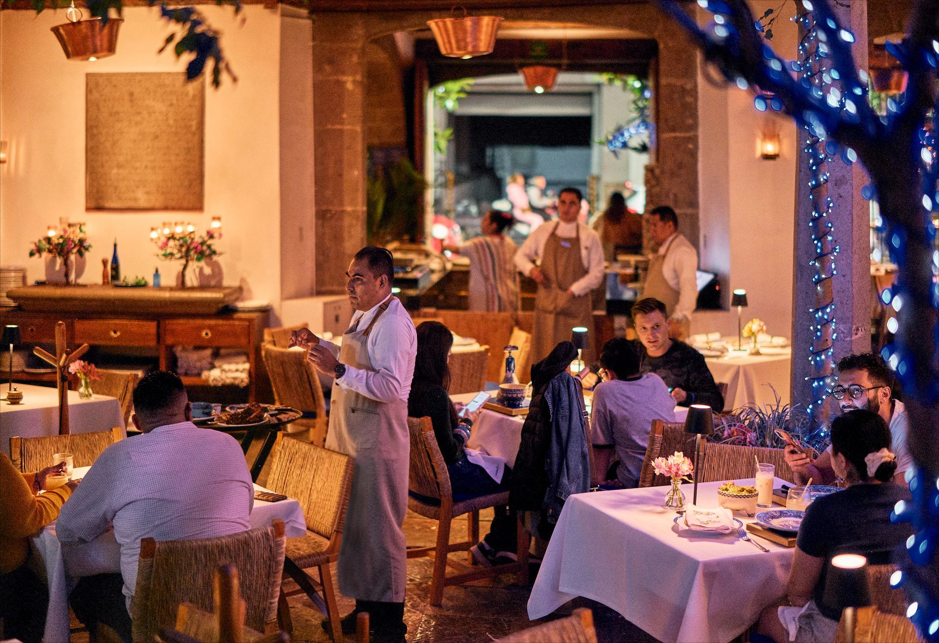 Patio de restaurante con iluminación tenue, comensales en mesas iluminadas con velas y luces azules en un árbol.