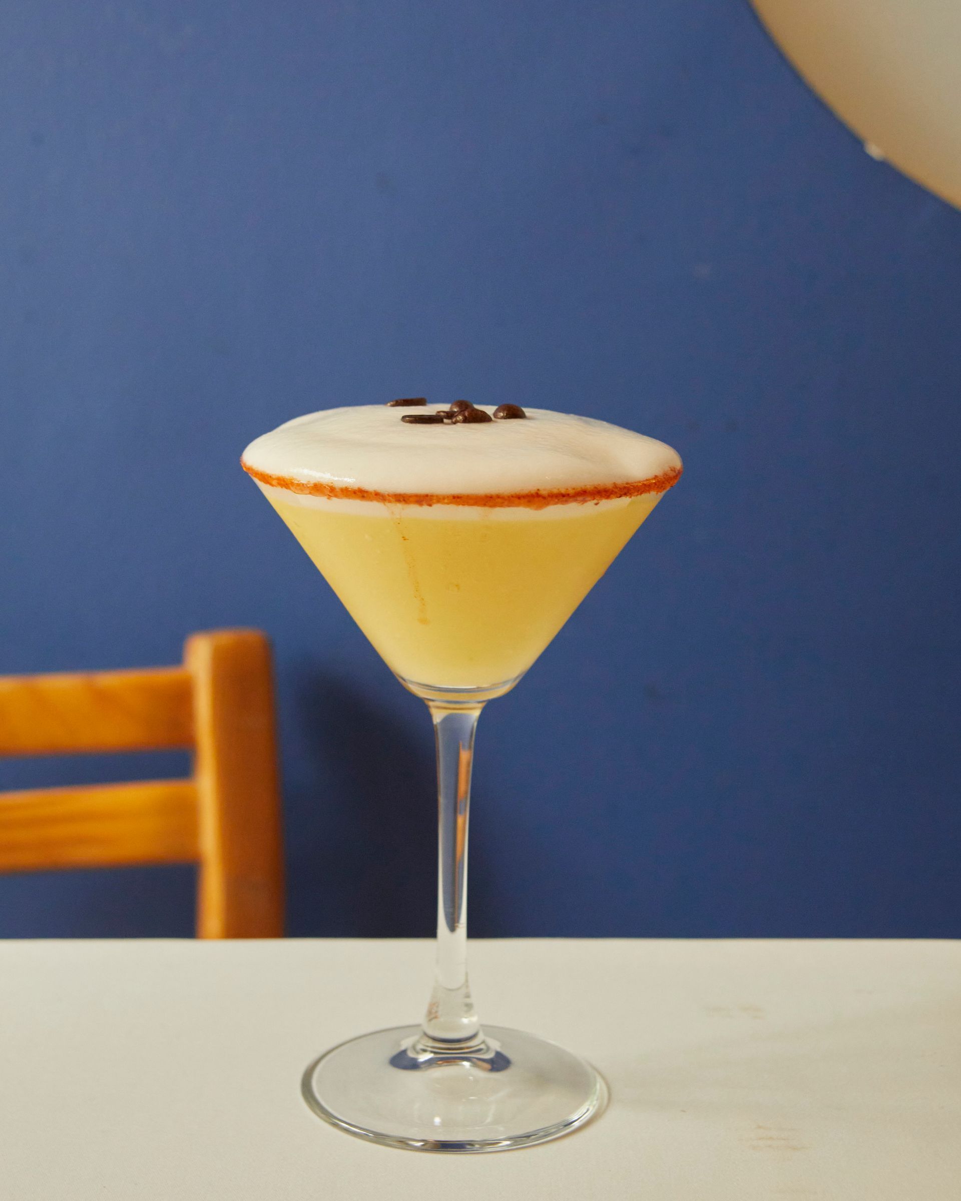Cóctel amarillo en una copa de martini con una capa blanca espumosa sobre una mesa con fondo azul.