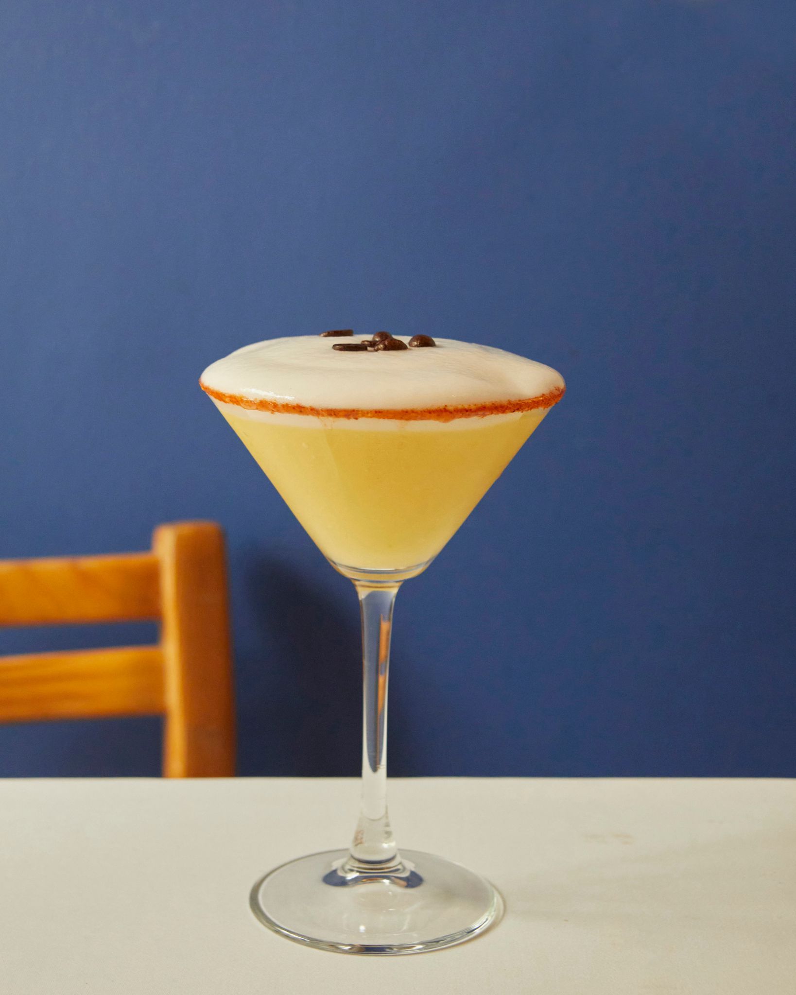 Una copa de martini con un cóctel amarillo pálido y espuma blanca sobre una mesa contra una pared azul.
