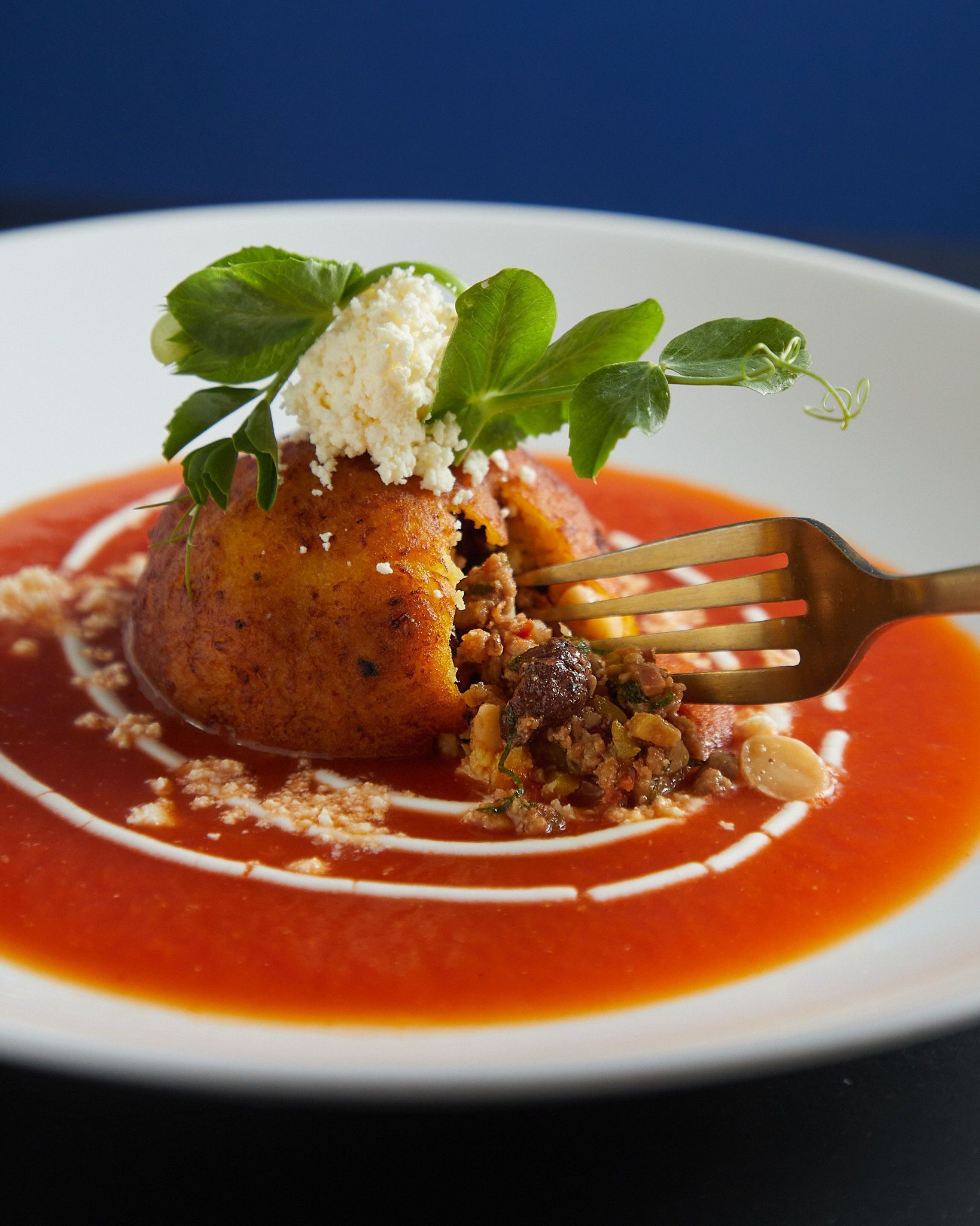 Tomar con un tenedor un tomate relleno en salsa roja, cubierto con albahaca fresca y queso rallado en un plato blanco.