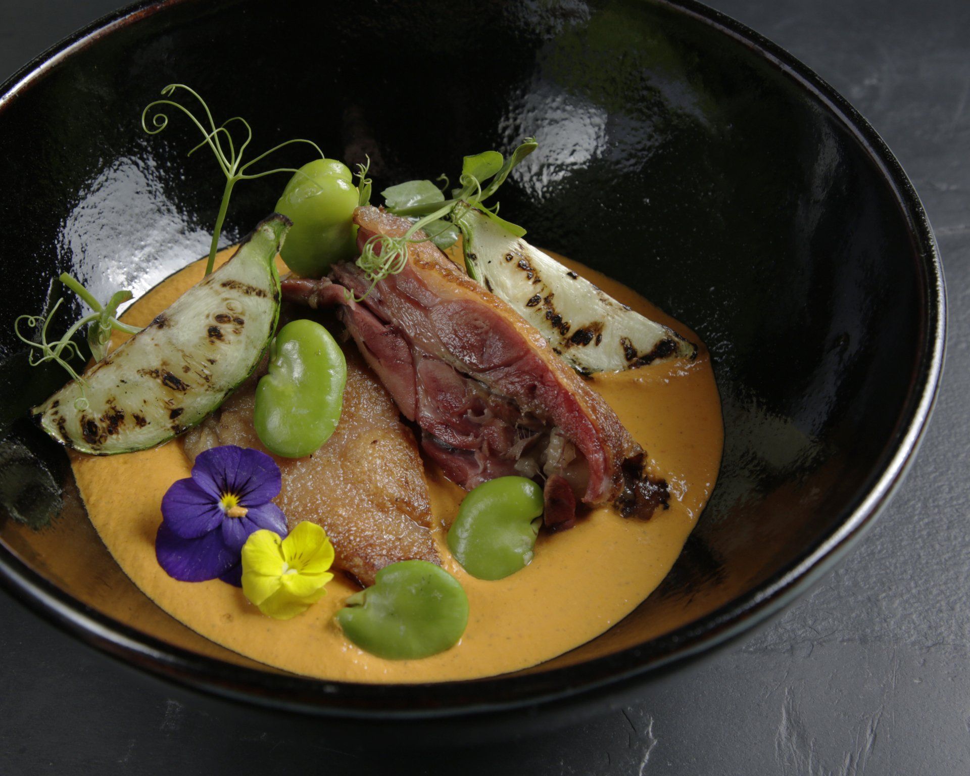 Plato gourmet servido en un cuenco negro con salsa, carne sellada, uvas verdes y flores comestibles.