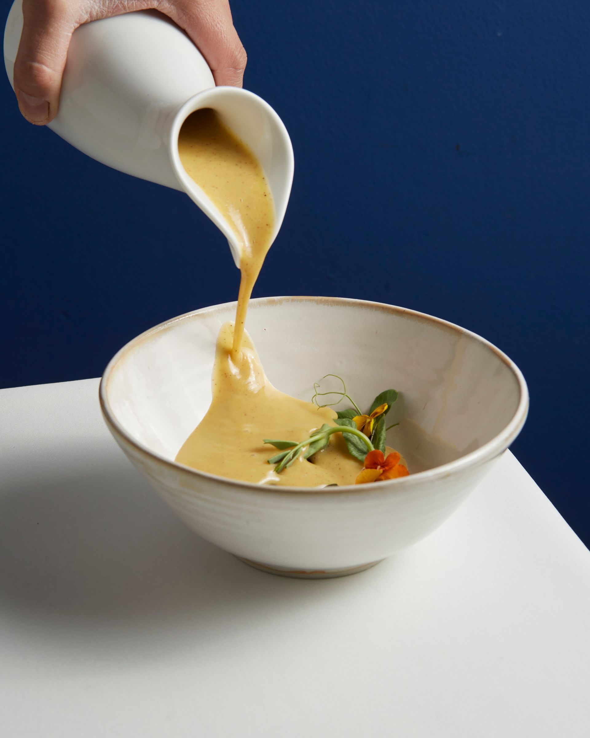 Sopa cremosa siendo vertida de una jarra blanca a un tazón sobre una mesa.