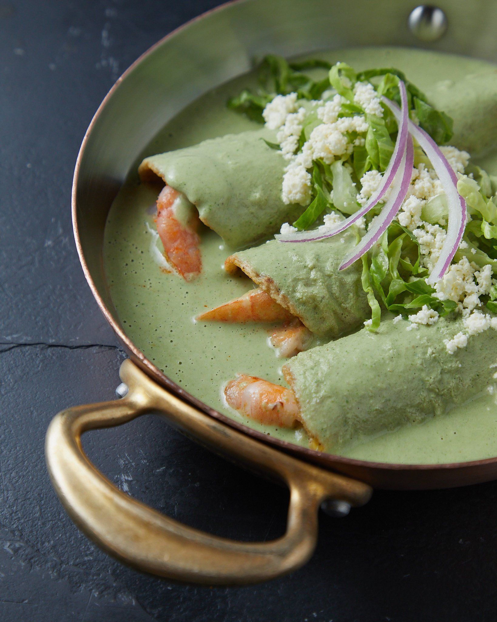 Crepes verdes enrolladas en una sartén con mango de latón, cubiertas con cebolla, hierbas y queso desmenuzado.