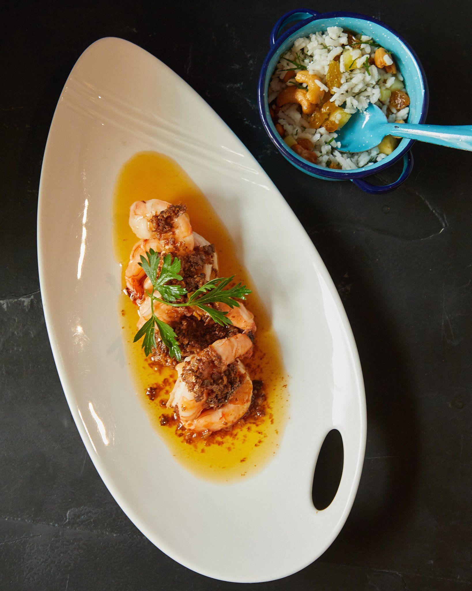 Pasta emplatada en salsa de naranja con hierbas, servida junto a un pequeño cuenco de arroz sobre una mesa oscura.
