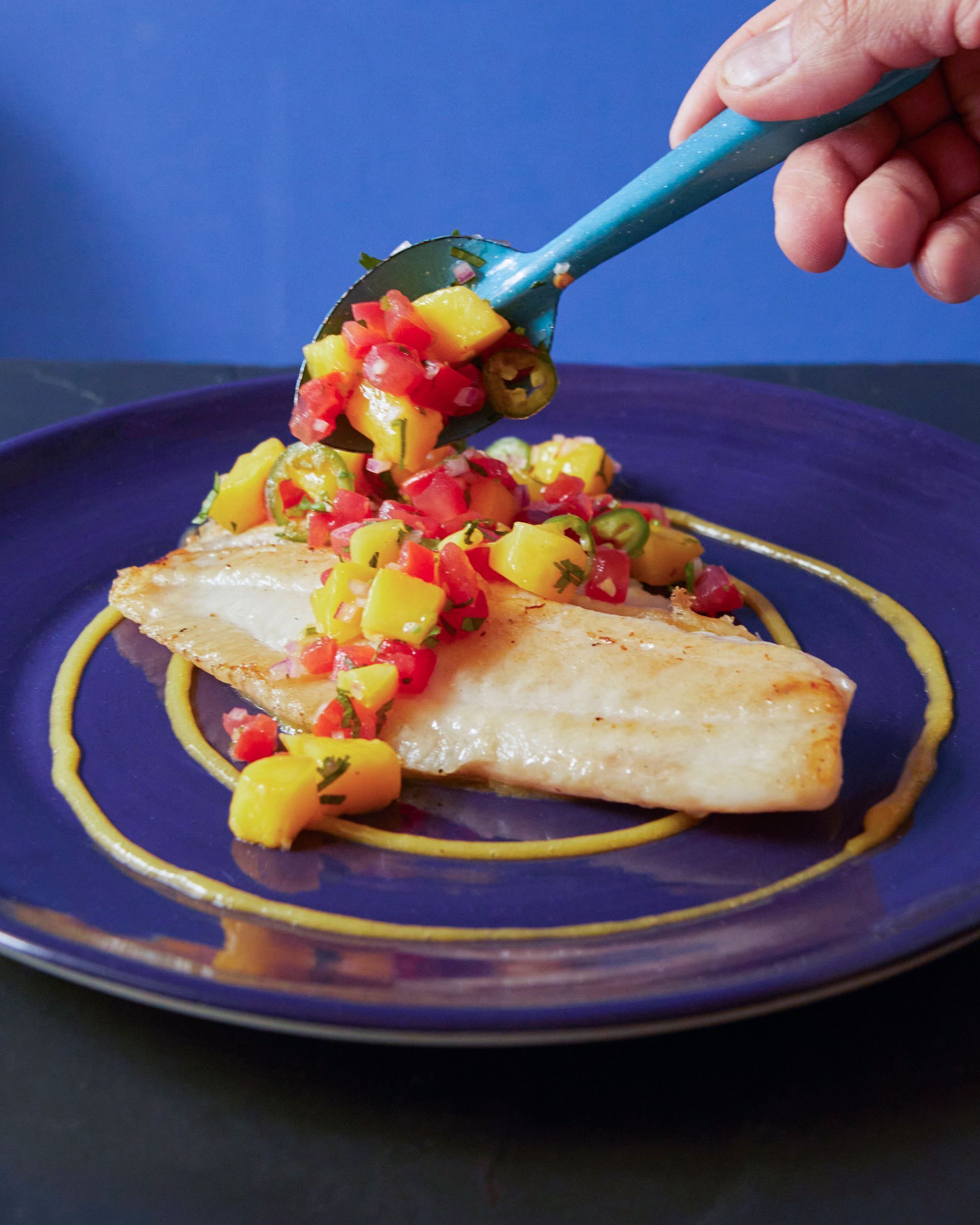 Filete de pescado en un plato azul cubierto con salsa de fruta picada, con una cuchara para añadir más salsa.