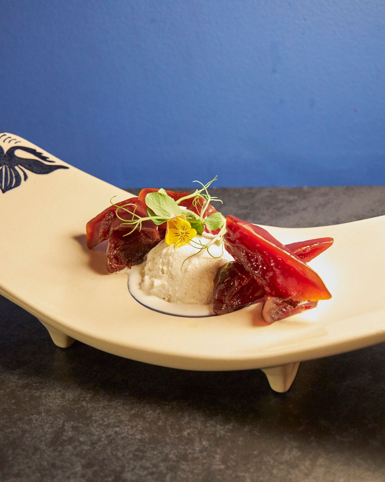 Postre emplatado con crema blanca, salsa de frutos rojos y menta en un plato curvo de crema sobre un fondo azul.