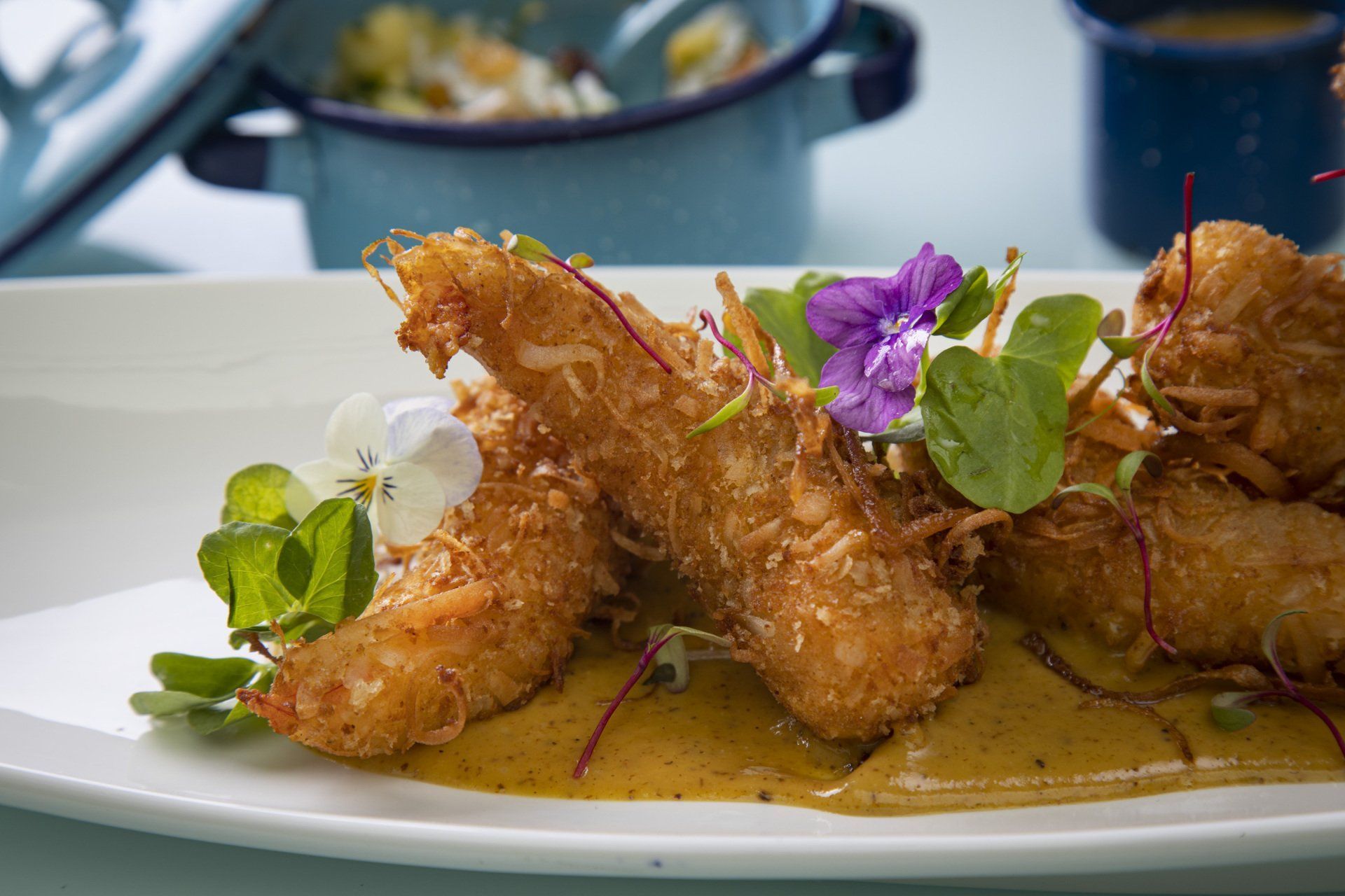 Gambas fritas emplatadas, adornadas con hierbas y flores moradas sobre un plato blanco.