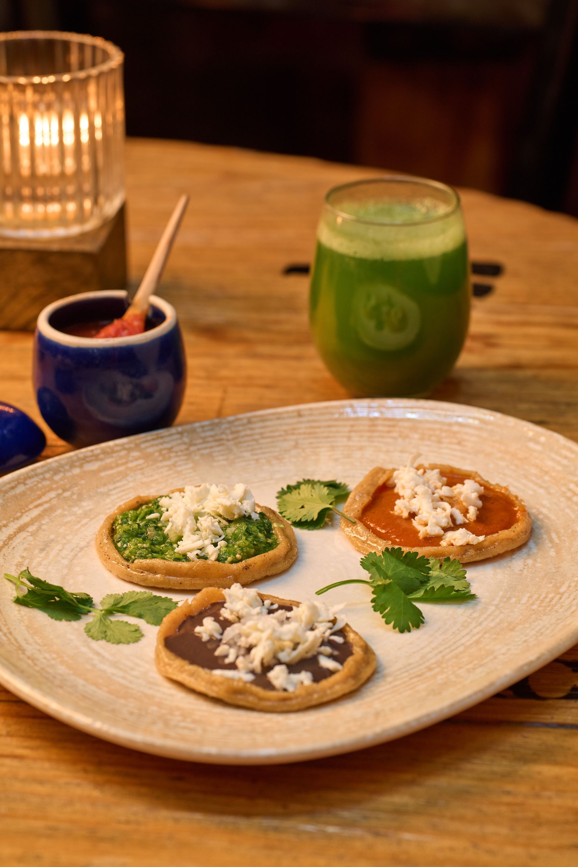 Tres pequeños pasteles en un plato con coberturas verdes, blancas y naranjas junto a las bebidas en una mesa de madera.