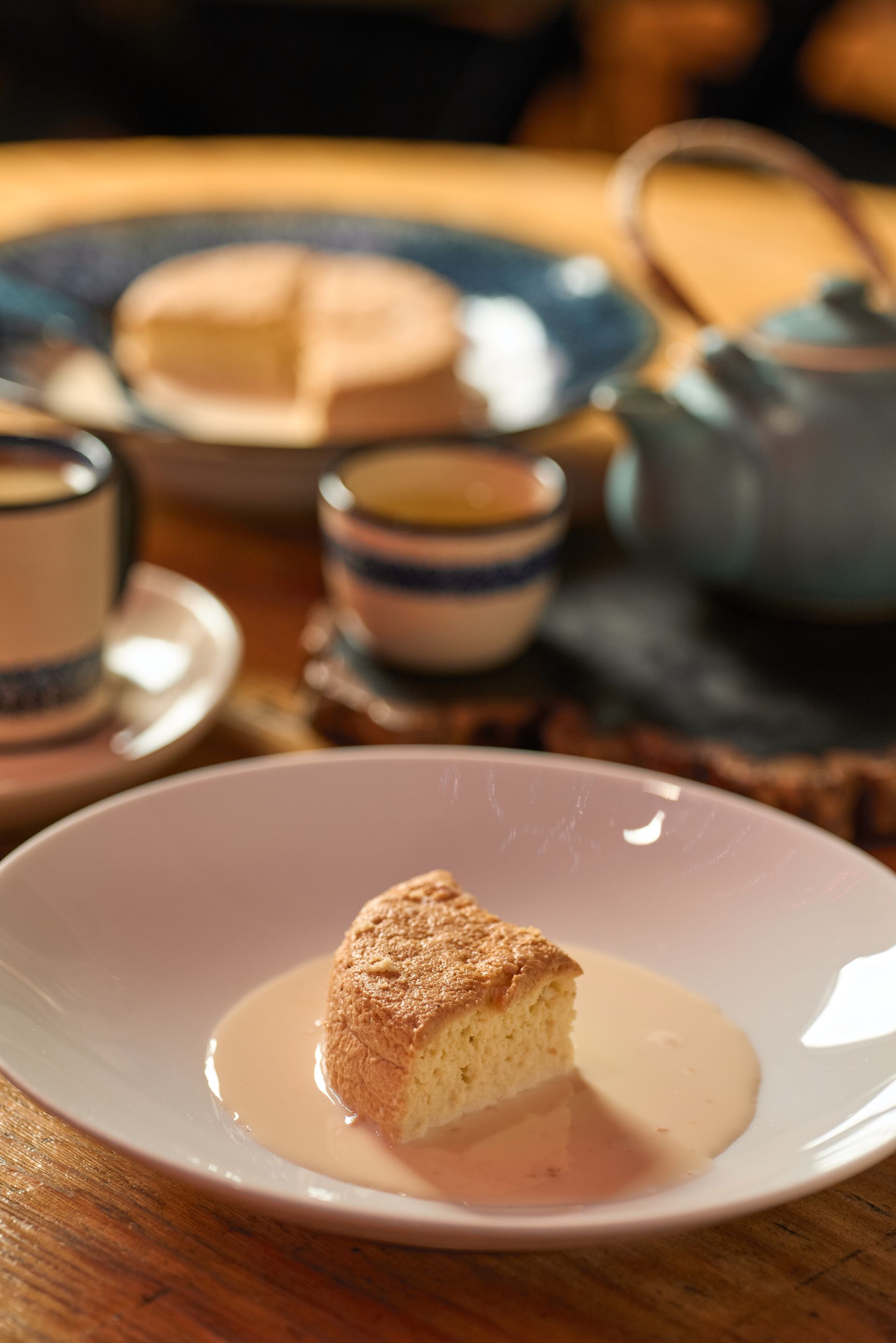 Una taza de té y una porción de pastel en un plato blanco con una tetera al fondo.