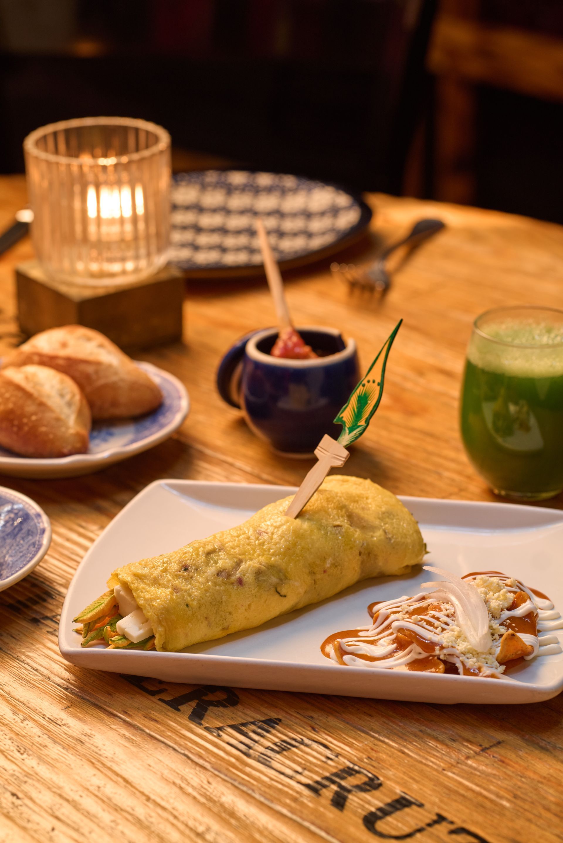 Mesa de brunch con una crepa amarilla, pan, salsas y jugo verde sobre una mesa de madera.