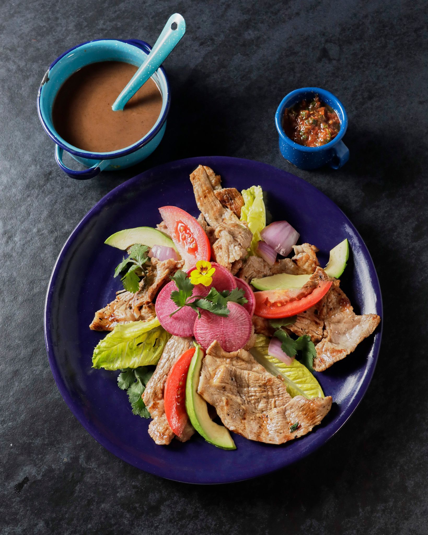 Ensalada de carne a la parrilla y aguacate en un plato azul con sopa y un pequeño cuenco de salsa en una mesa oscura.