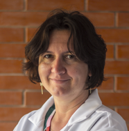Una mujer con una bata de laboratorio blanca sonríe frente a una pared de ladrillos.
