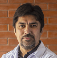Un hombre con barba y bata blanca está parado frente a una pared de ladrillos.