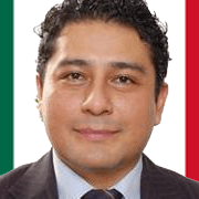 Un hombre con traje y corbata sonríe delante de una bandera mexicana.