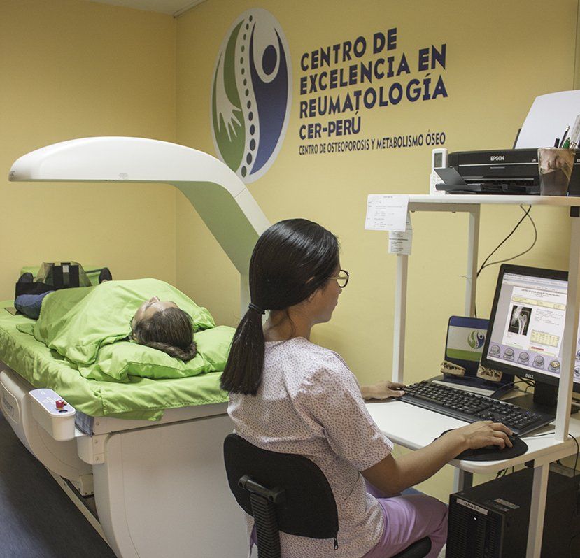 Una mujer está sentada en un escritorio con una computadora frente a un cartel que dice centro de excelencia en reumatología