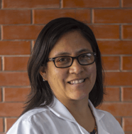 Una mujer que lleva gafas y una bata blanca sonríe delante de una pared de ladrillos.