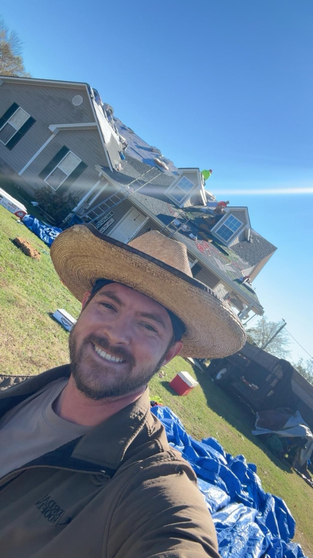 Man smiling outdoors in a large straw hat, with a house and blue sky behind him.