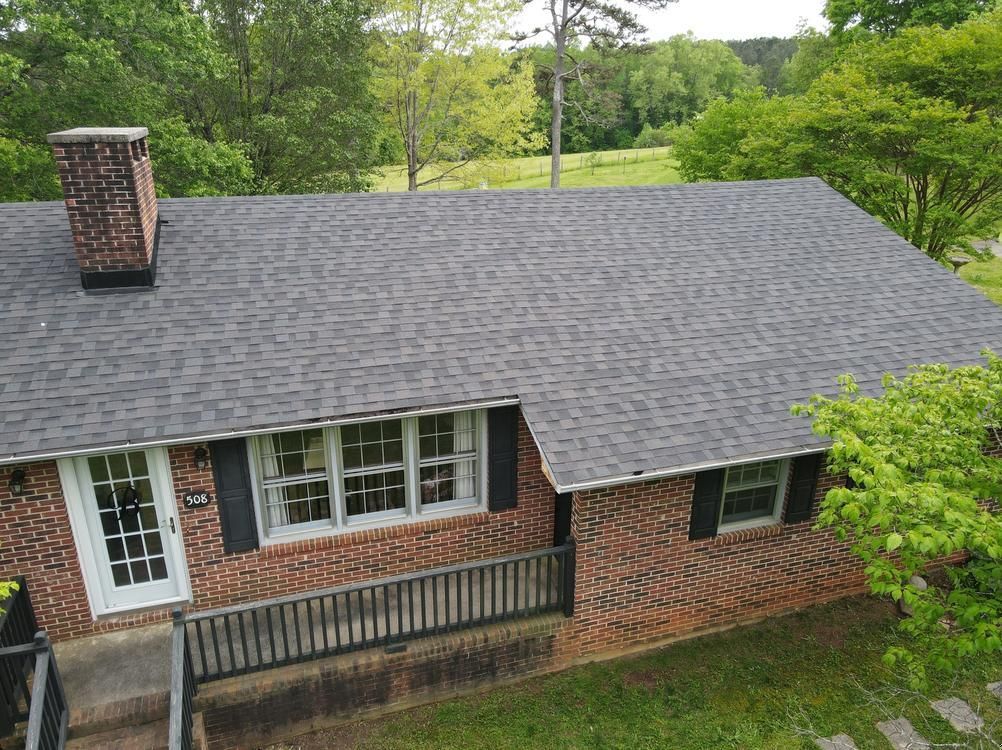 Brick house with gray roof and chimney, surrounded by trees and greenery.